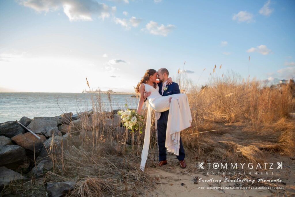 A couple in wedding attire kiss on a sandy, grassy beach at sunset, with rocks and water in the background.