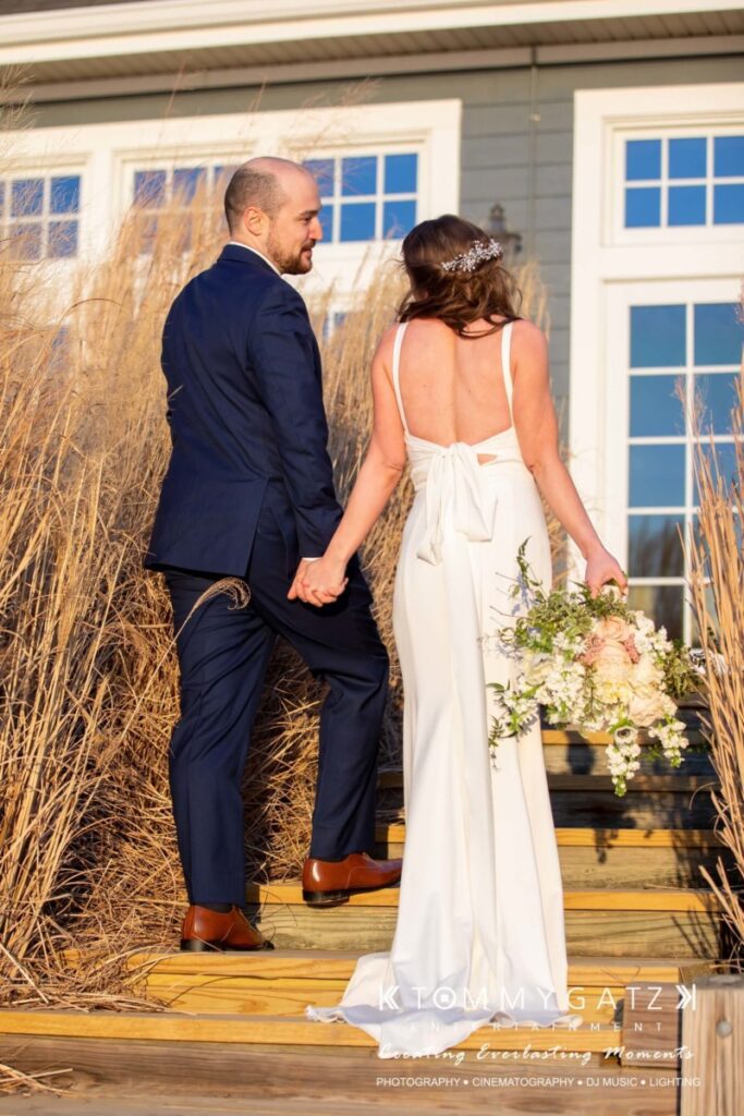 A bride and groom holding hands, walking up wooden steps outdoors, surrounded by tall grasses. The bride holds a bouquet and wears a white gown; the groom is in a navy suit.