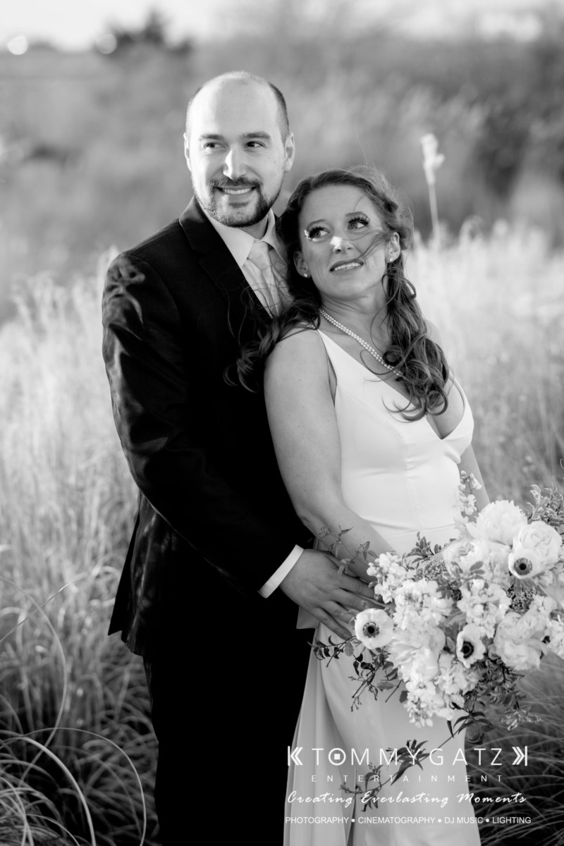 A couple dressed in wedding attire stand in a field with tall grass. The man is slightly behind the woman, who is holding a bouquet of flowers.