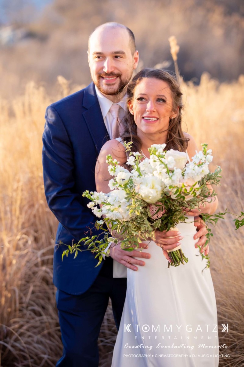A couple in formal attire stand in a field. The woman holds a bouquet of white flowers, while the man stands behind her, arms around her waist. They both look toward the distance.