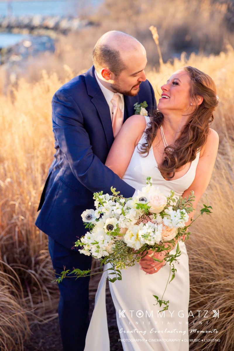Bride and groom smiling at each other, standing in front of tall grass, with the bride holding a bouquet of white flowers.