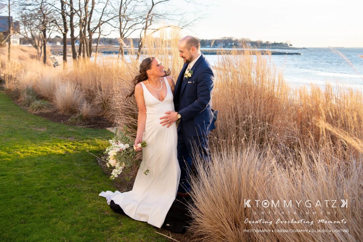 A bride and groom stand smiling by a lakeside, surrounded by tall grass. The bride holds a bouquet in one hand and her gown's train in the other as the groom rests his hand on her waist.