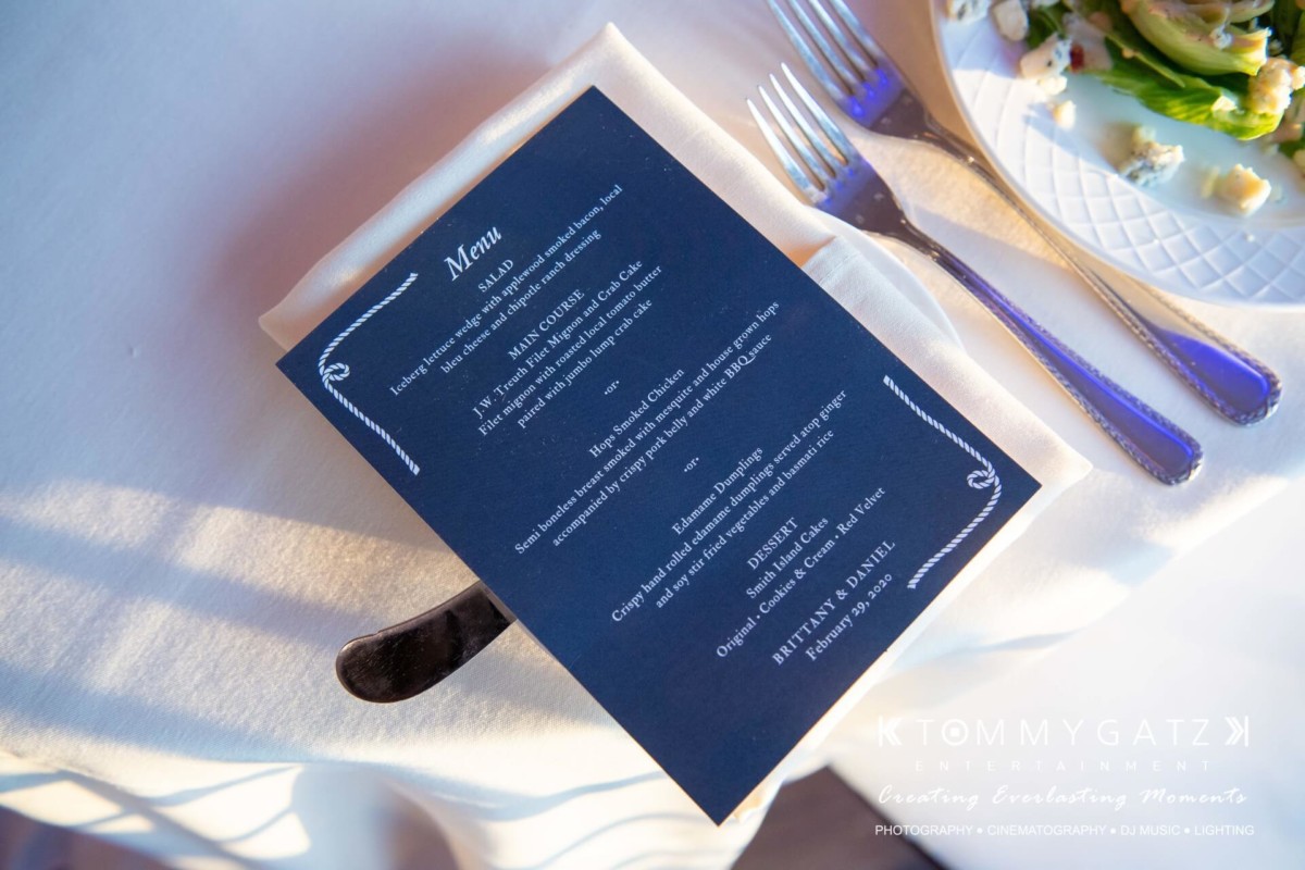 Dark blue menu on a white plate with silverware, set beside a plate of salad and white flowers.