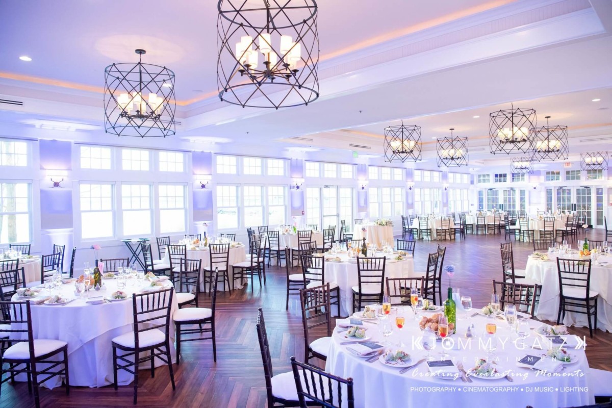 Elegant banquet hall with round tables set for an event. White tablecloths, wooden chairs, and chandeliers create a formal atmosphere. Large windows line the walls, allowing natural light in.