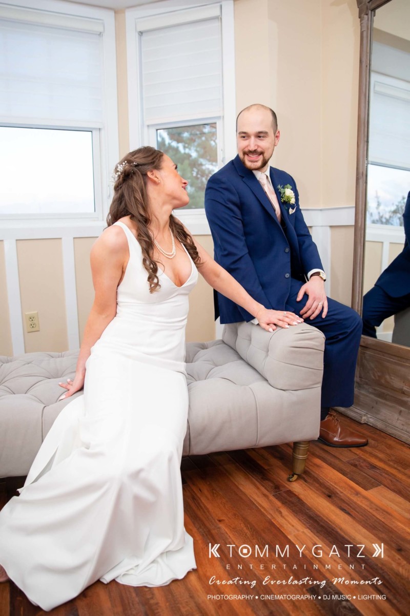 A bride in a white dress and a man in a suit sit on an upholstered bench, smiling at each other near a large mirror.