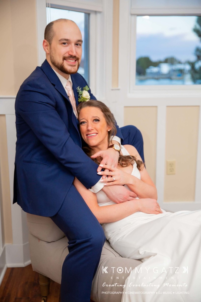 A couple, dressed in formal attire, sits together on a cushioned chair in a bright room. They are smiling at the camera.