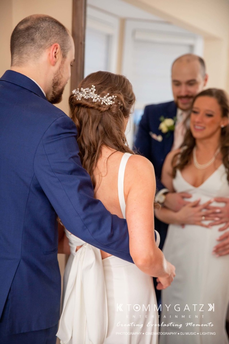 A bride and groom stand closely together in front of a mirror. Another man in a suit stands nearby, partially visible. The bride wears a white dress with a decorative hairpiece.