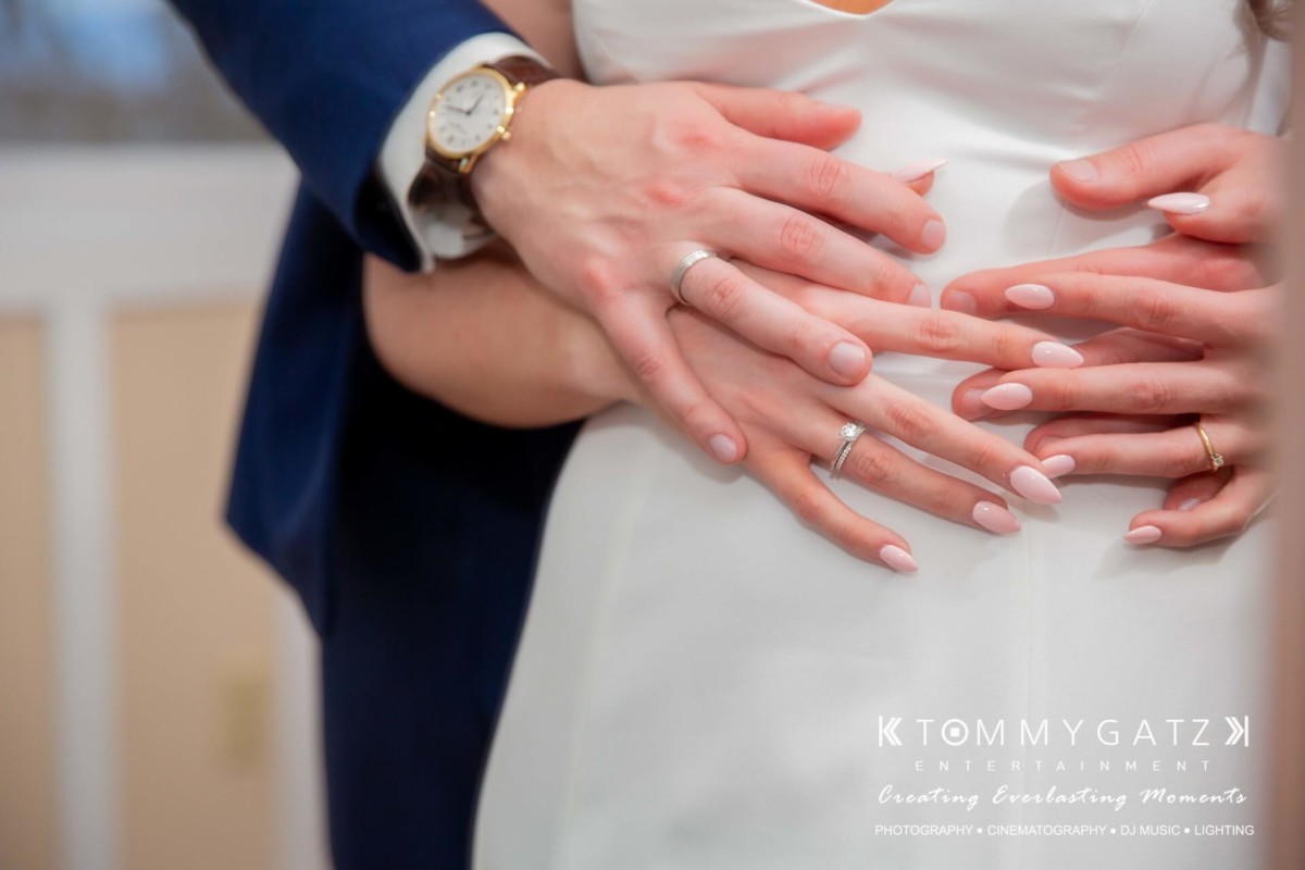 Close-up of a couple embracing, showing hands with wedding rings. The man's hand supports the woman's waist, over a white dress, showcasing a watch and a suit sleeve.