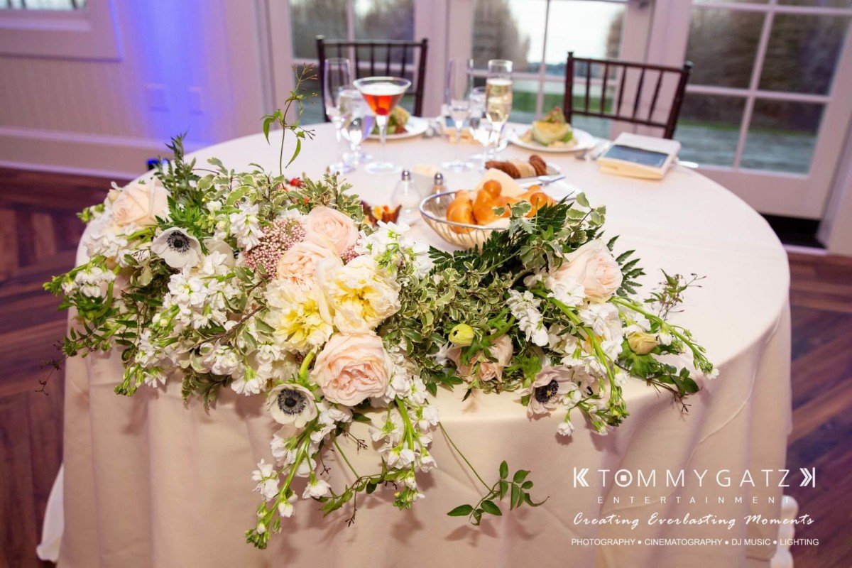 Elegant table setting with a white tablecloth, floral centerpiece, and various dishes, including bread and drinks, at an indoor event with natural light coming through the windows.