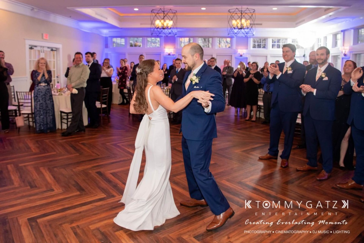 A bride and groom dance in a reception hall as guests look on and clap. The groom is in a blue suit, and the bride in a white dress.