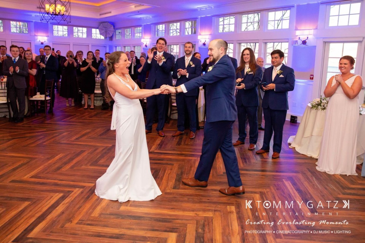 A bride and groom dance in a room as guests applaud. Elegant decor and wooden floor are visible, with large windows in the background.
