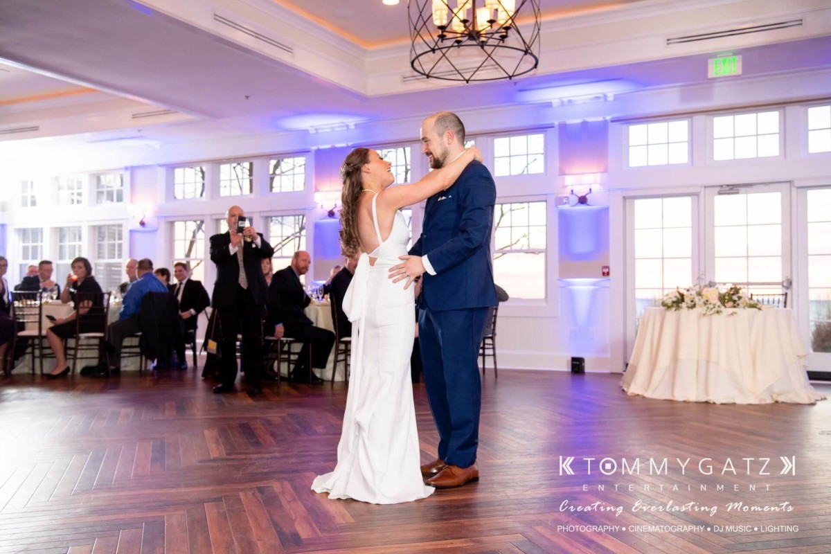 A bride and groom share their first dance on a wooden floor in a bright reception hall, with seated guests in the background.