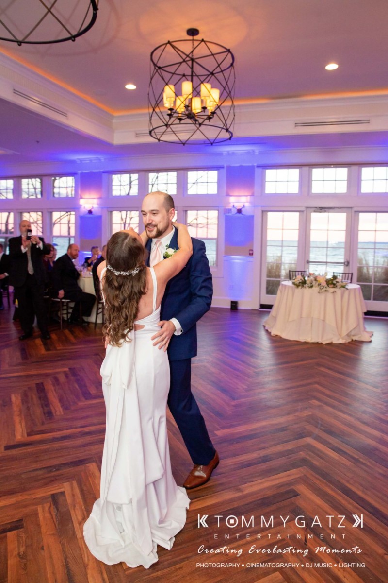 A couple dances in a decorated ballroom. The woman is in a white dress, and the man is in a suit. Guests are seated at tables in the background.