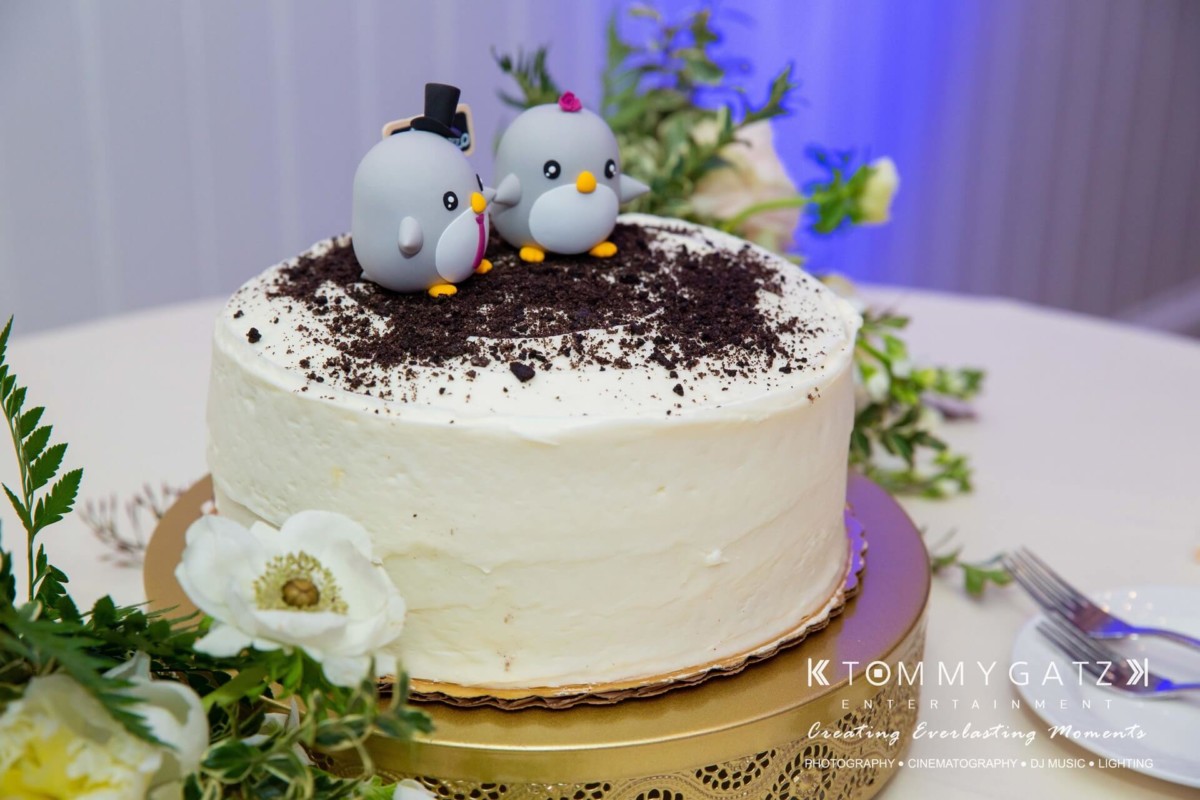 A white cake topped with two penguin figurines, one wearing a top hat, on a table with flowers and a cake server.