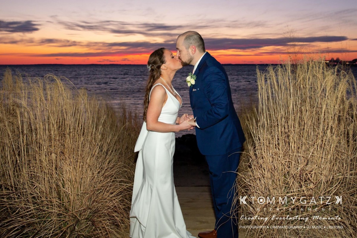 A bride and groom kiss at a beach during sunset, surrounded by tall grasses.