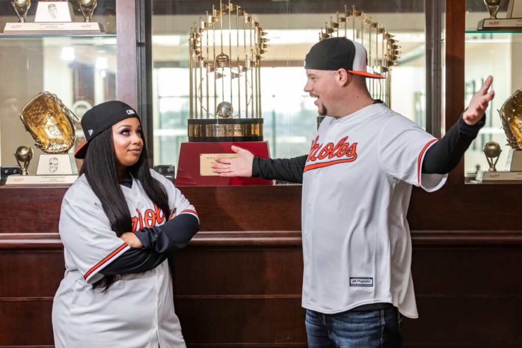 Two people in matching baseball jerseys stand in front of a trophy display case. One person gestures widely, while the other crosses their arms.