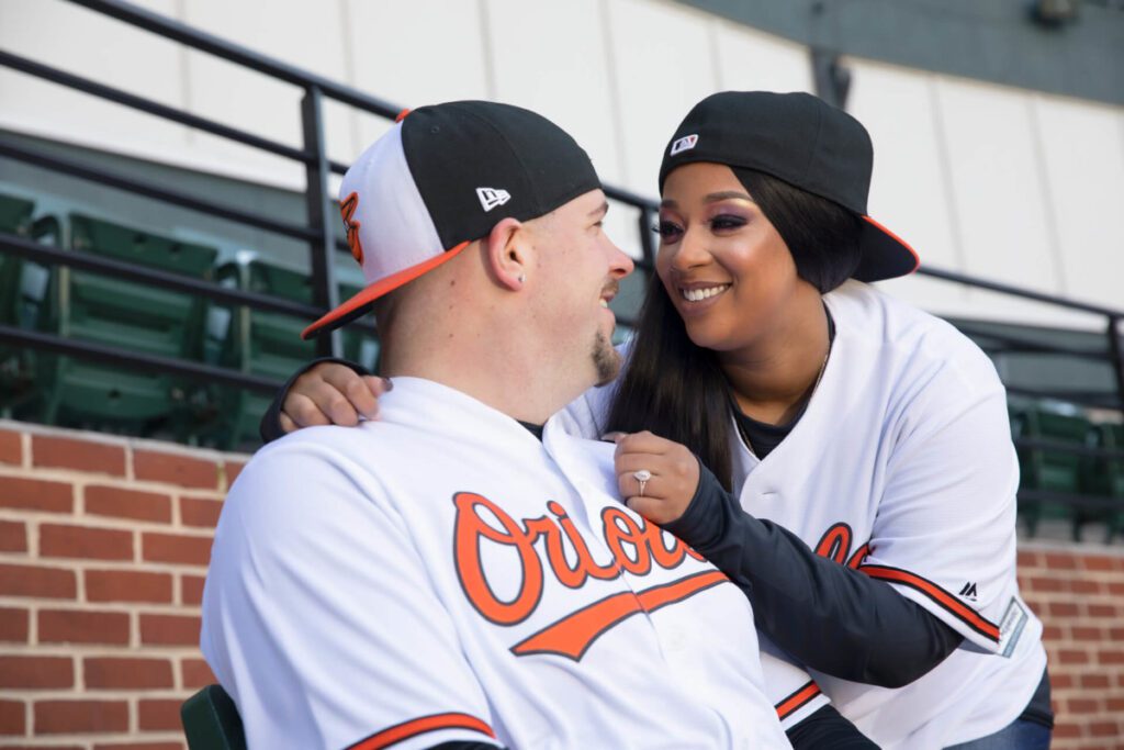 Two people in matching Orioles jerseys and caps, sitting together in a baseball stadium, smiling at each other.