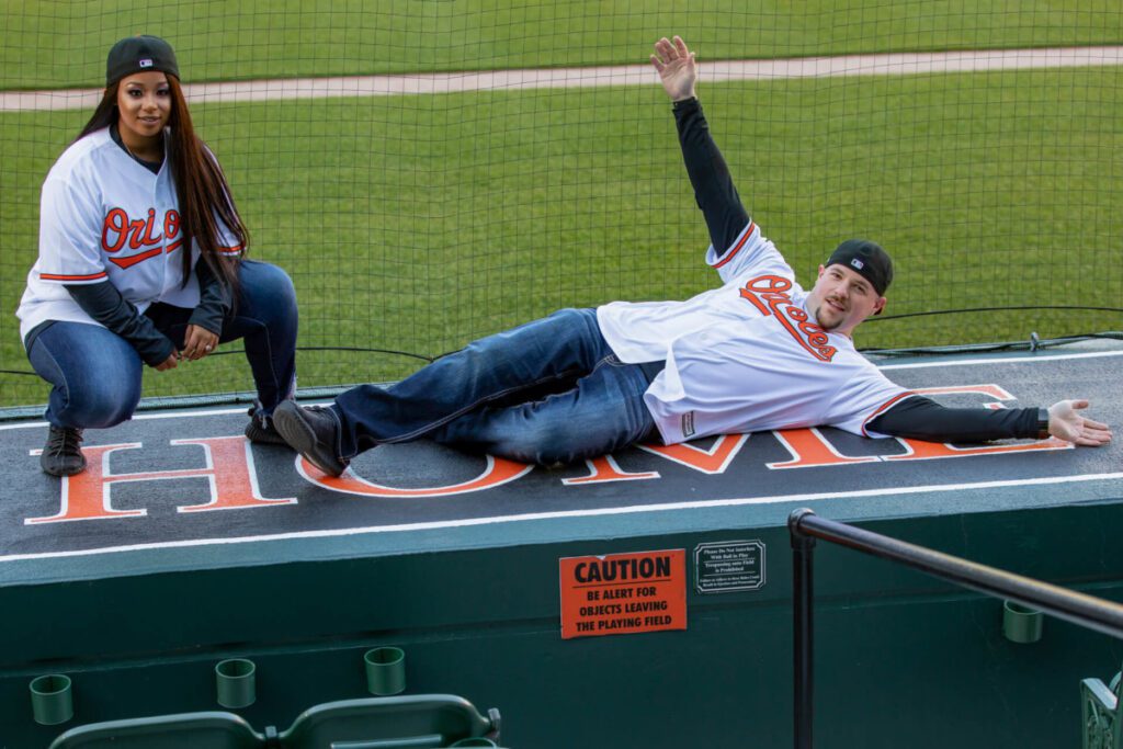 Two people in Orioles jerseys pose on the "HOME" dugout bench at a baseball field. One kneels, and the other is lying down with arms outstretched, both smiling.