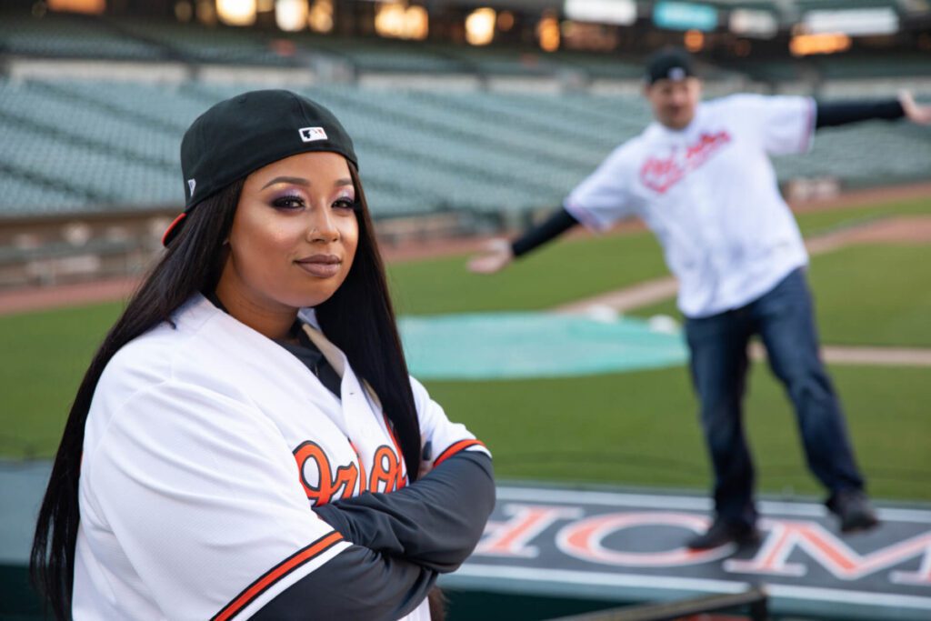 Two people in baseball jerseys on a field; one is posing confidently in the foreground, while the other playfully gestures in the background.