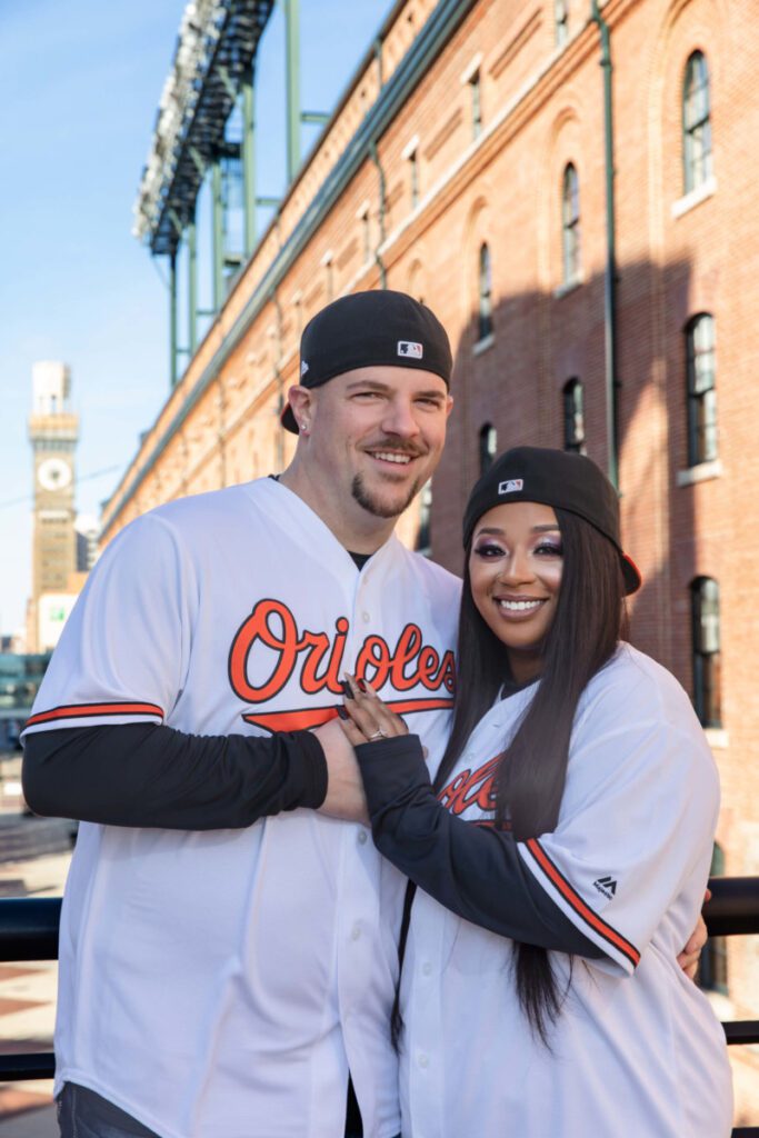A couple in matching baseball jerseys and caps stands smiling in front of a red brick building with a clock tower in the background.