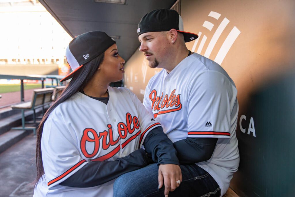 Two people wearing Orioles baseball jerseys and caps, sitting and looking at each other in a dugout.
