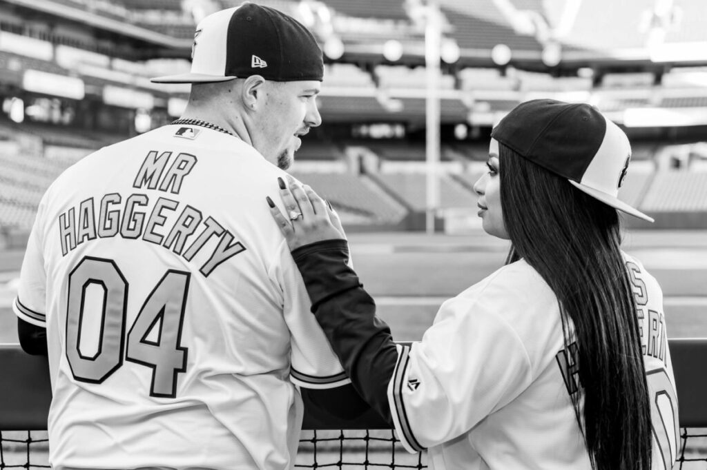 A couple in matching baseball jerseys stand at a sports stadium, with the back of their shirts reading "Mr. Haggerty 04" and "Mrs. Haggerty." They are looking at each other.