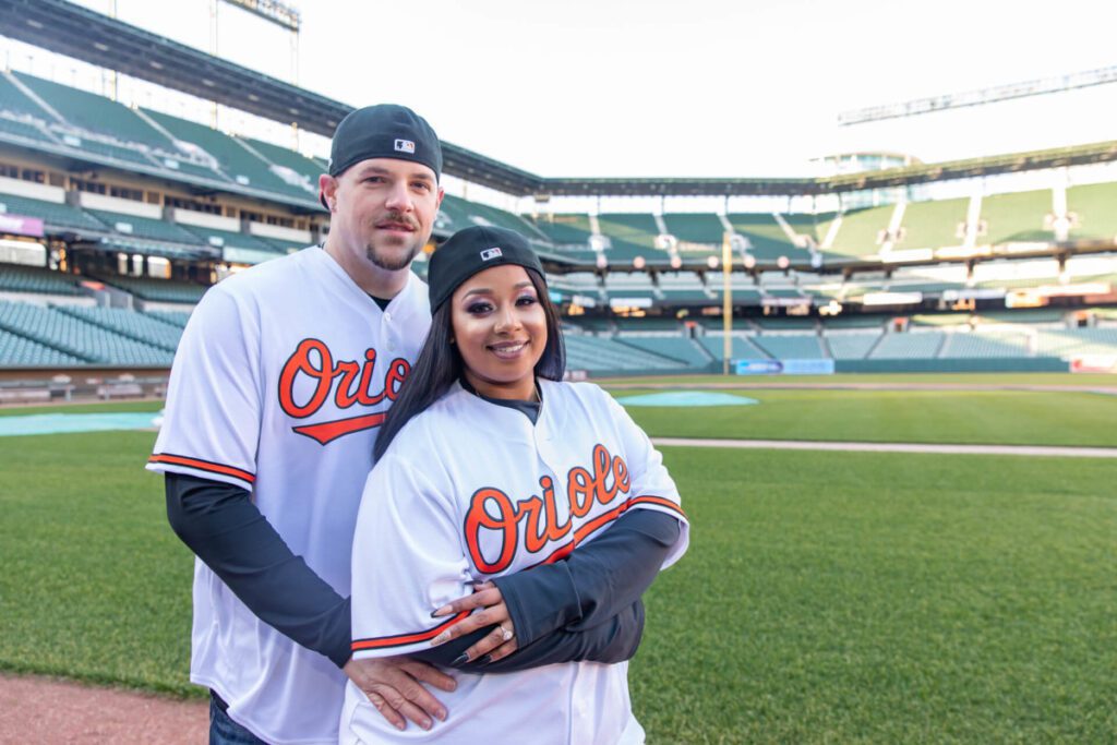 Two people in "Orioles" baseball jerseys stand on a green field in an empty stadium. The person in front wears a black cap.
