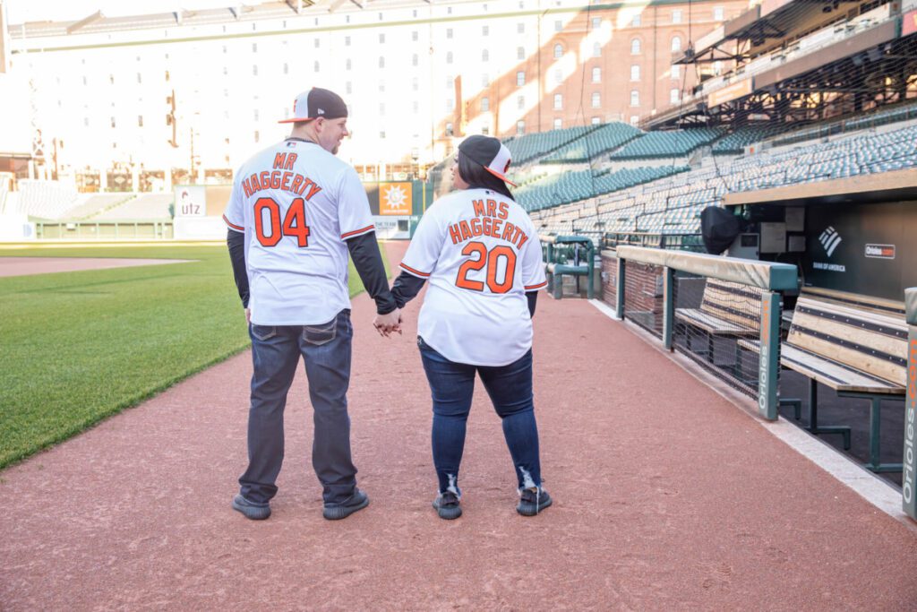 A couple in matching jerseys labeled "Mr. Haggerty 04" and "Mrs. Haggerty 20" holds hands, walking through a baseball stadium.