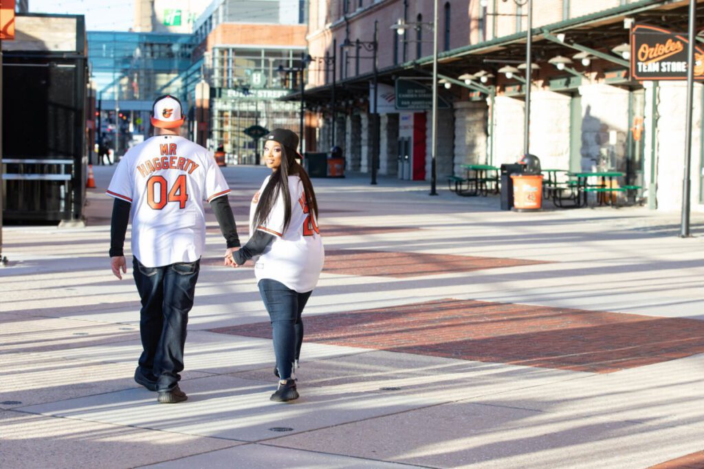 A couple wearing matching baseball jerseys walks hand-in-hand along a sunlit street near a team store.