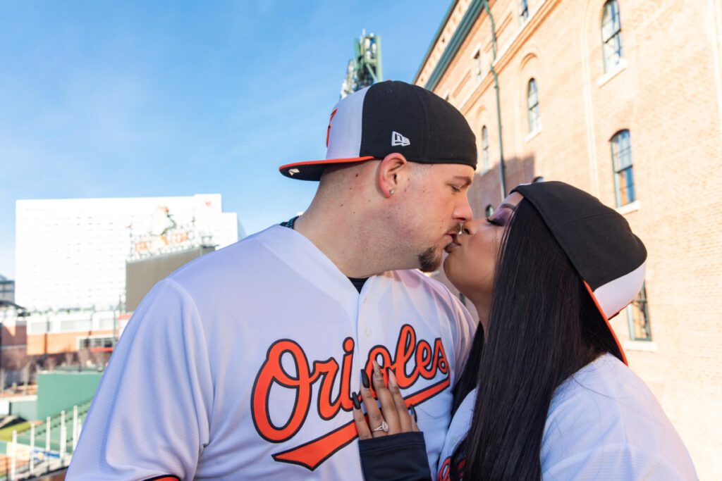 A couple in Orioles jerseys kisses outside a baseball stadium.