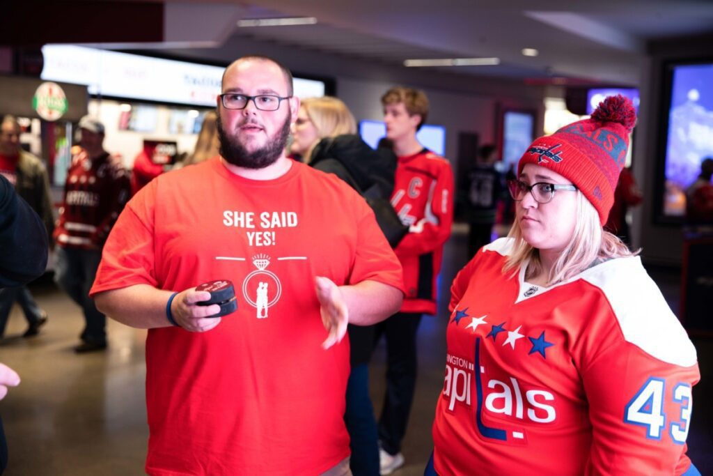 Two people in hockey attire inside an arena; one wears a "She said yes!" shirt, holding a puck, and the other wears a Washington Capitals jersey.