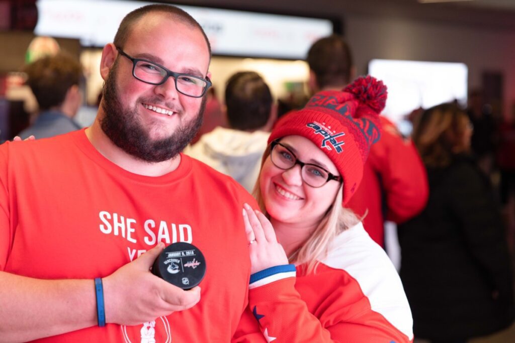 A man and woman smile, holding a hockey puck. Both wear red shirts; the woman has a red hat. People are in the background.