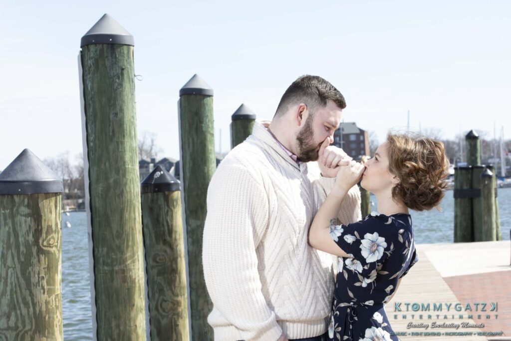 A couple stands by a waterfront, holding hands and leaning close. The man wears a cream sweater and the woman wears a floral dress. Dock posts and water are in the background.