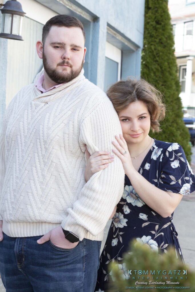 A man in a white sweater and jeans stands next to a woman in a floral dress, who is leaning on his arm outside a building.