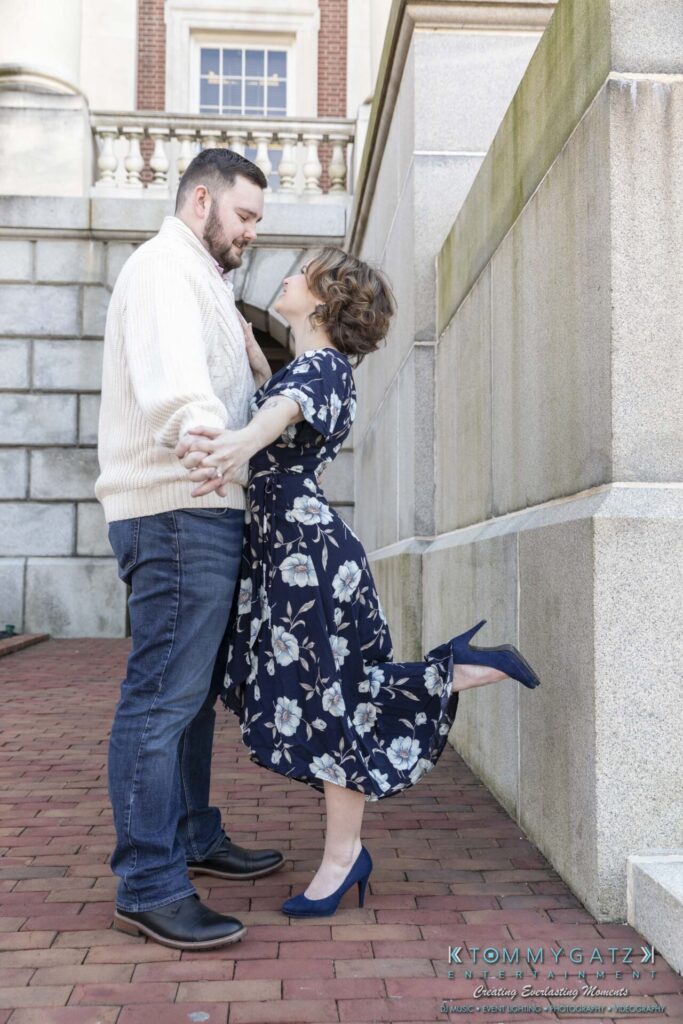 A couple poses affectionately near a brick wall. The woman in a blue floral dress holds the man's hands and lifts one leg. He wears a cream sweater and jeans.