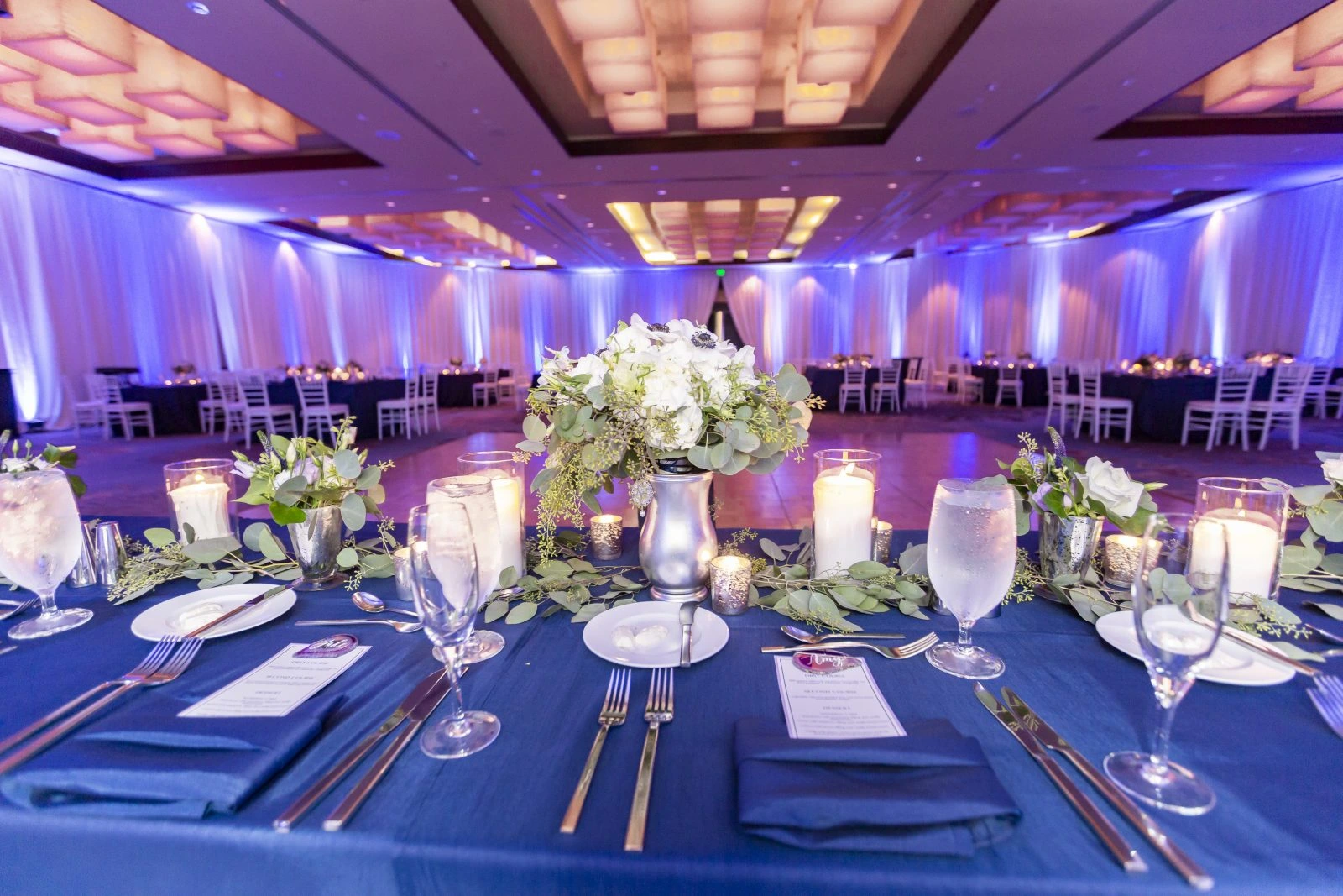 080418-Amy-Jake-Non-Watermark-8 - TOMMY GATZ ENTERTAINMENT Elegant banquet table set with blue linens, glassware, candles, floral centerpiece, and menus in a spacious room with mood lighting.
