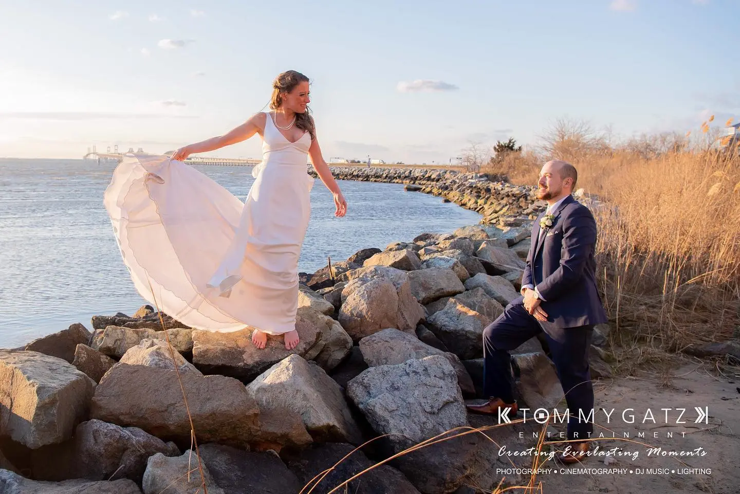 A bride in a white dress and a groom in a suit pose on rocks by the water under a clear sky.