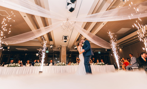 A couple shares a first dance at a wedding reception, surrounded by sparklers and guests seated at a long table in an elegant venue with a draped ceiling.