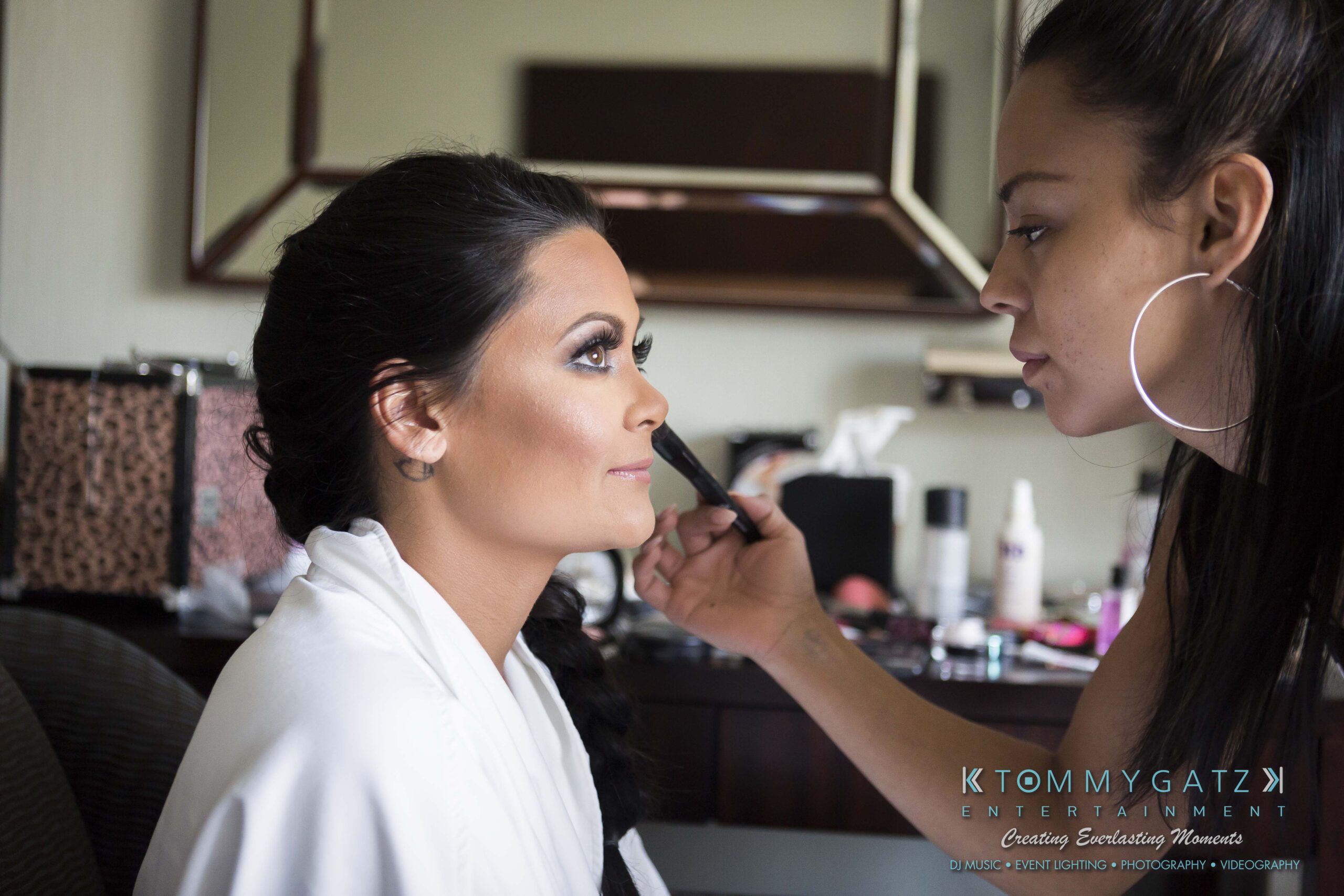 A makeup artist applies eyeliner to a woman sitting in a chair, with beauty products scattered on a table in the background.