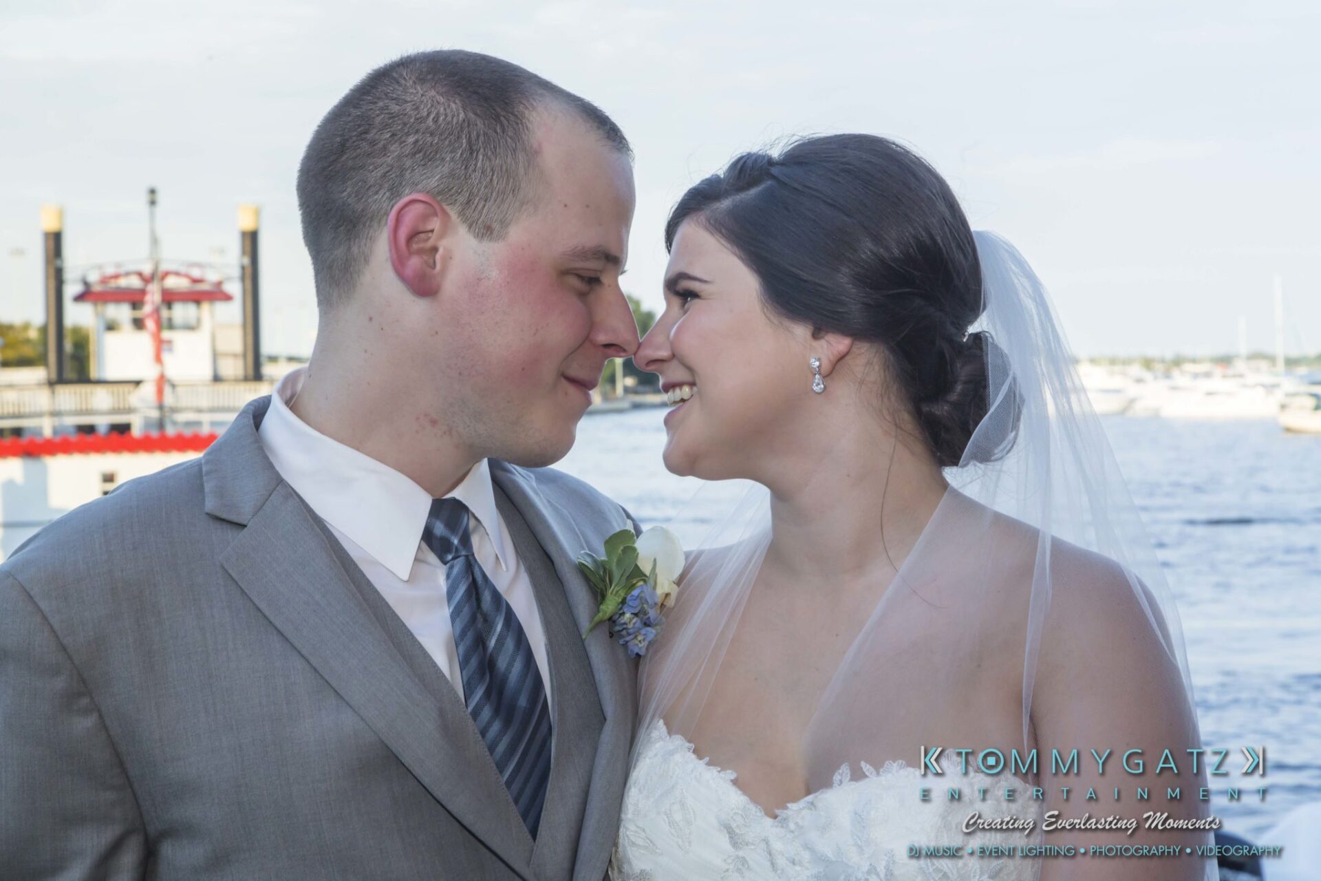 A bride and groom smile at each other, standing near a waterfront.
