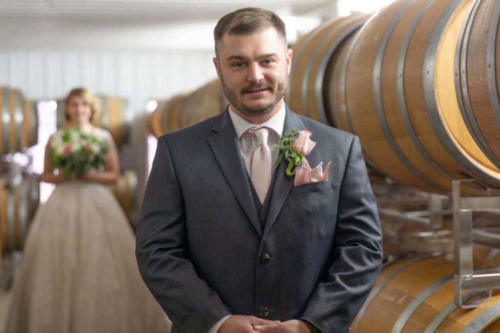 A groom in a suit stands in front of wine barrels, with a bride holding a bouquet in the background.