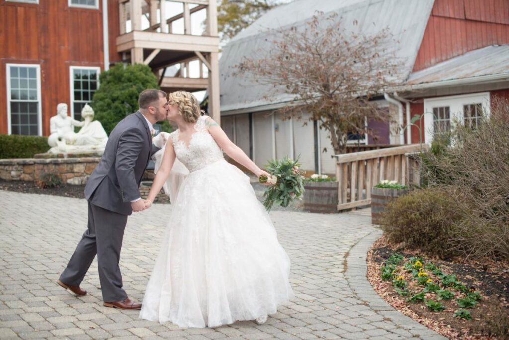 A bride and groom kiss while holding hands on a cobblestone pathway in front of rustic buildings and a small garden. The bride holds a bouquet.