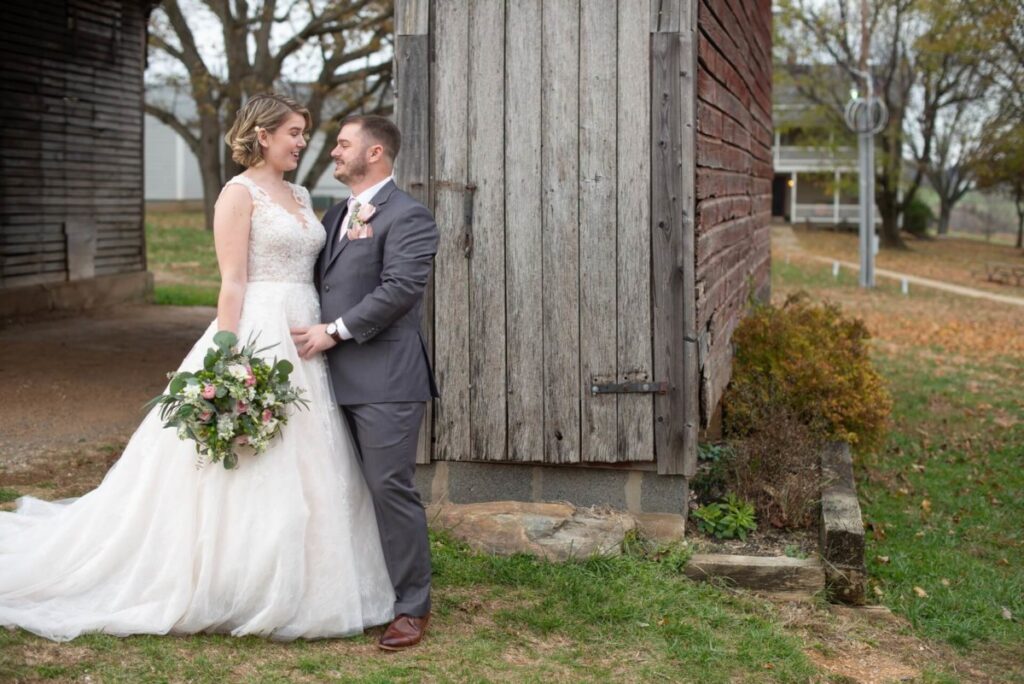 A bride and groom stand together outdoors by a rustic wooden building. The bride holds a bouquet and wears a white dress, and the groom wears a gray suit.