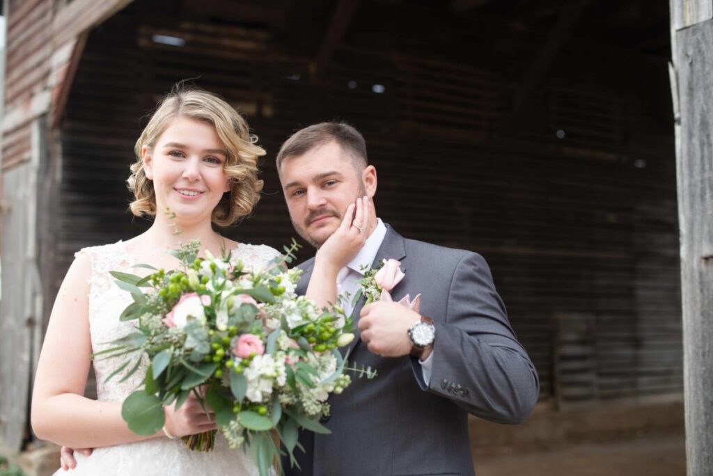 A bride and groom pose for a photo in front of a rustic wooden structure. The bride holds a bouquet of flowers, and the groom touches her face while wearing a suit with a boutonniere.