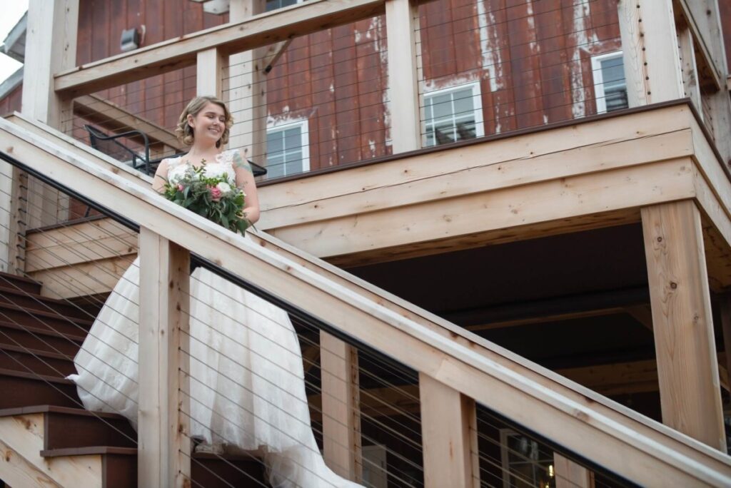 A woman in a white wedding dress holds a bouquet while descending wooden stairs of a rustic building.