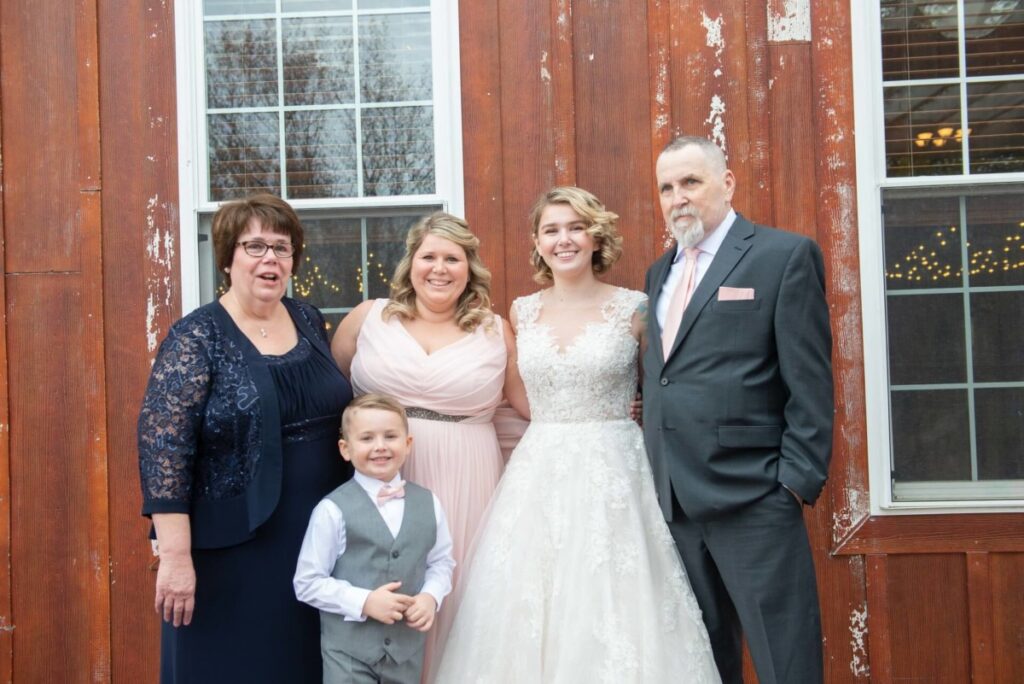 Five people pose for a group photo in formal attire in front of a wooden building with windows.
