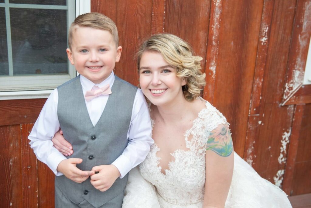 A young woman in a white lace dress and a child in a gray vest with a bow tie smile in front of a wooden wall.