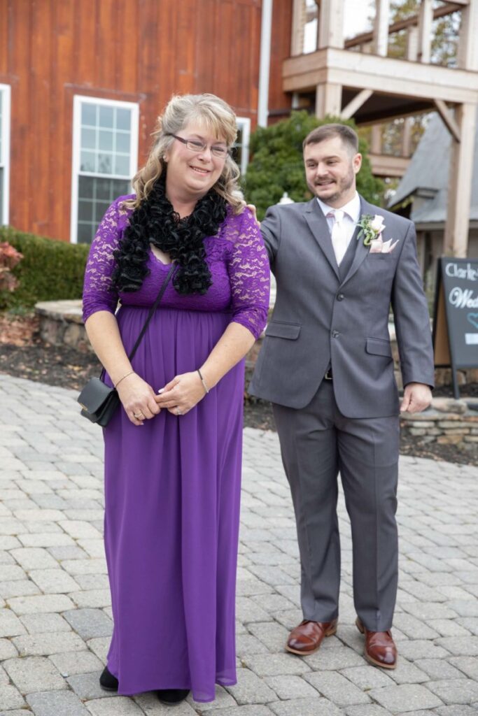 A woman in a purple lace dress and a man in a gray suit stand outdoors on a paved walkway near a wooden building.