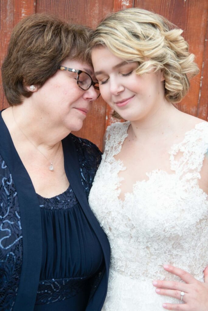 Two women standing closely, with eyes closed, in front of a wooden background. One is in a lace dress, the other in a blue outfit. They appear to share a tender moment.
