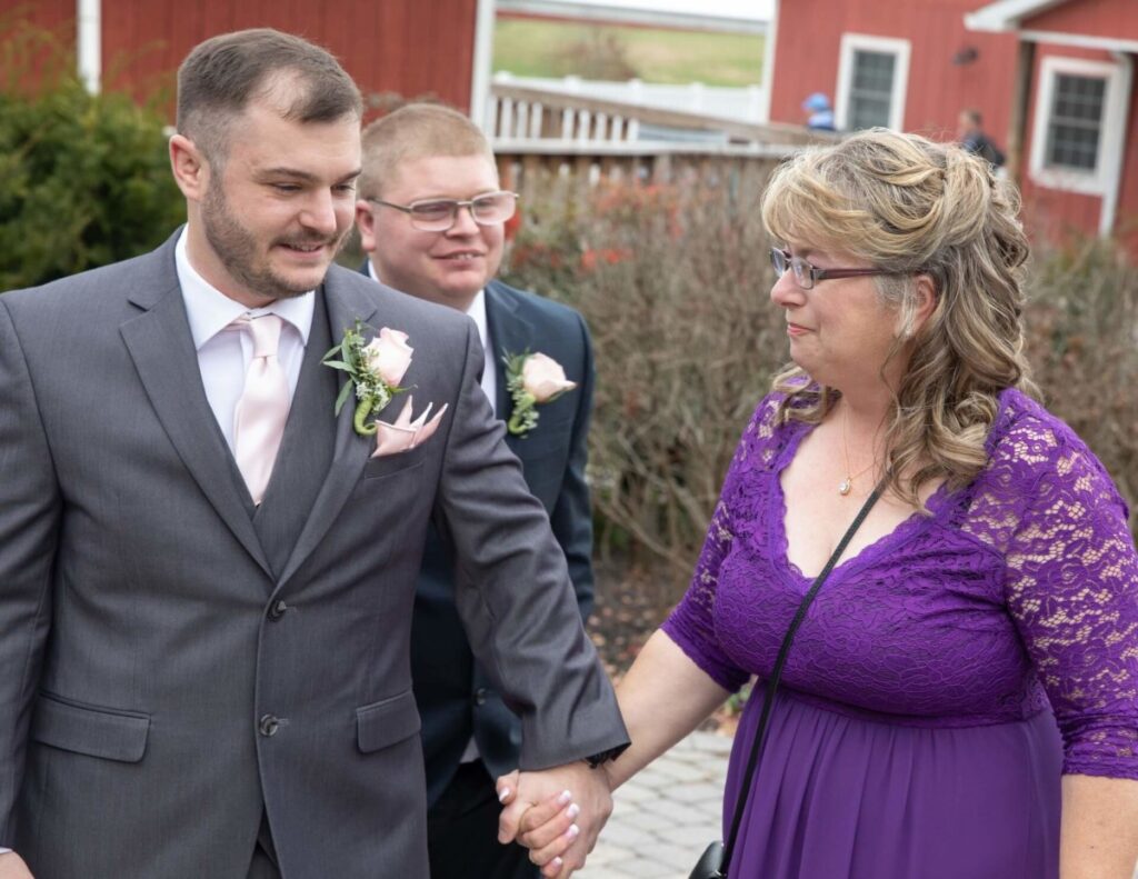 A man in a suit holding hands with a woman in a purple dress outdoors. Another man in a suit is in the background. They stand on a paved walkway with red buildings and shrubs nearby.